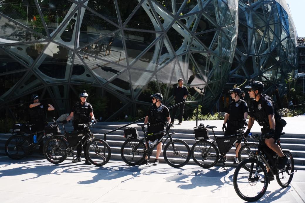 Five Seattle police wearing helmets and cameras on their heads stand next to their bikes outside the Amazon spheres on a sunny day. Aside from two people walking down the stairs nearby, they appear to be alone.