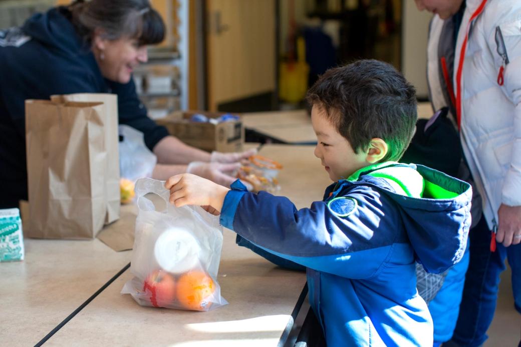 A five-year-old student wearing a big blue jacket grabs a plastic bag with an apple, an orange and a white container visible inside. There are brown sack lunches in the background and school staff handing more food out to the rest of the line behind the student.