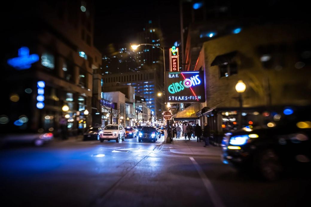 A night shot of a street. There are cars and neon lights. A sign on a building says "Gibsons"