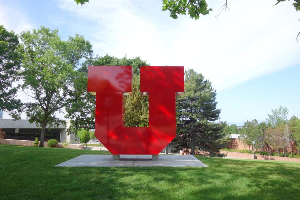 A large red sculpture of the University of Utah’s "block U” logo stands on a square concrete pad in a sunny grassy area. 