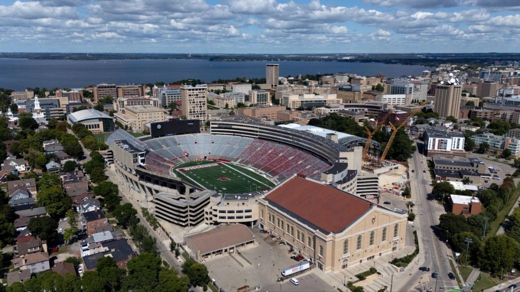 An overhead view of the UW Madison campus with building and a football field and surrounding neighborhoods. Water can be seen in the distance.