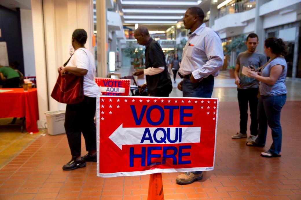 People standing in a line behind a red sign that says “Vote Aqui Here.”