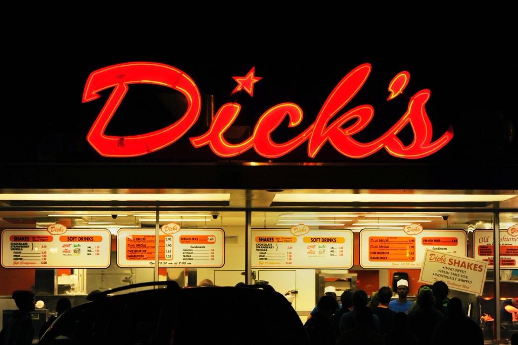 An image of Dick’s Drive-in at night, with the logo lit up and below it an illuminated window and ordering window]