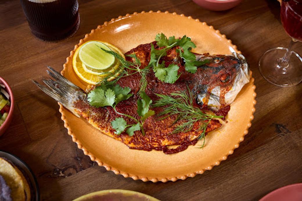 An orange plate sits on a wooden table. A cooked fish topped with sauce, herbs, and lemon slices rests on the plate. 