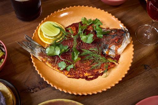 An orange plate sits on a wooden table. A cooked fish topped with sauce, herbs, and lemon slices rests on the plate.