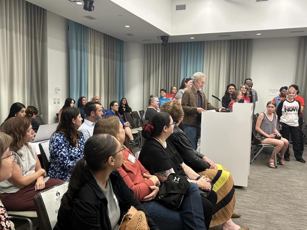 A crowded community meeting room where a man speaks at a white podium while a diverse audience of adults listens from rows of chairs. Several attendees wear advocacy shirts, including one reading 'Respect Now.'"