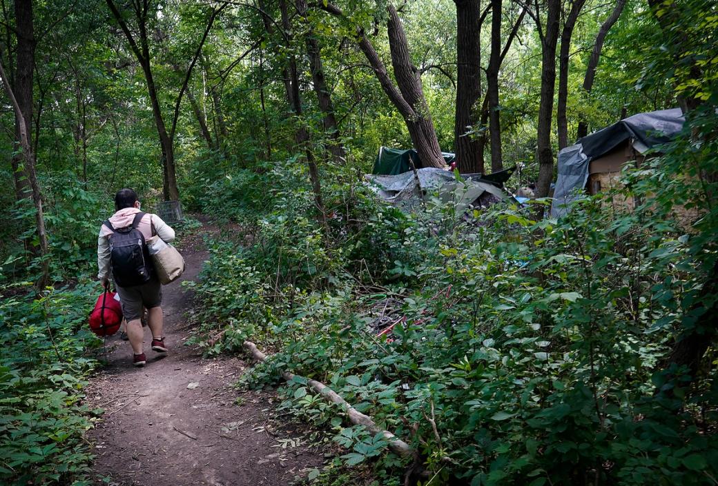 Person wearing backpack and carrying bags walking through the woods on a well worn path, off to the right is a few makeshift tents