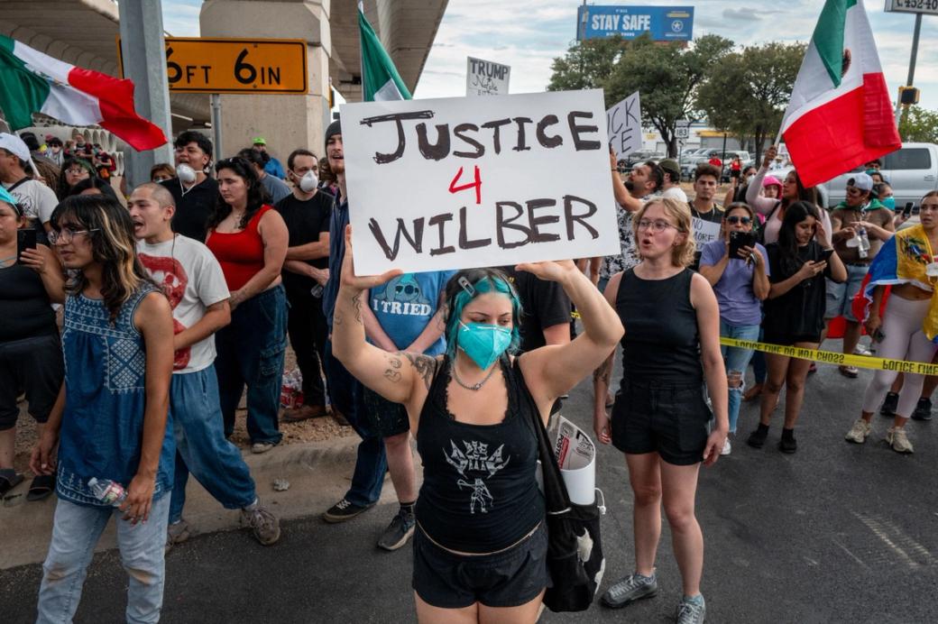 A person wearing a blue face mask and black tank top carries a white sign that says “Justice 4 Wilbur” on it.