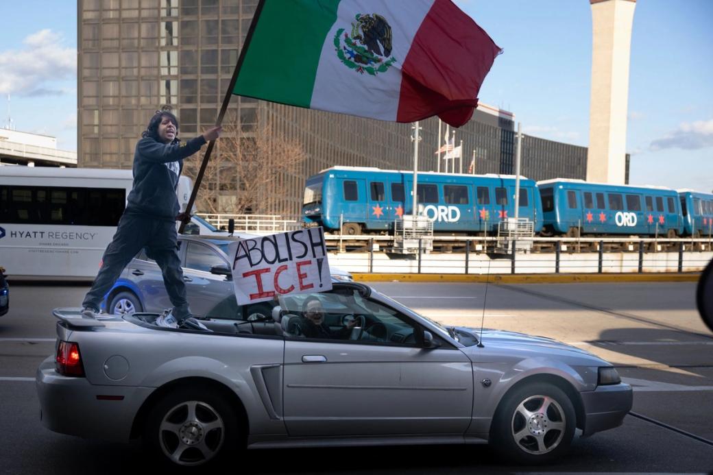A person standing on a silver car holding a Mexican flag. The driver of the car is holding a sign that says "Abolish ICE"