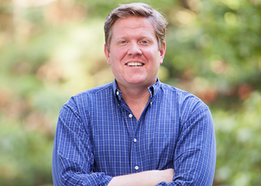 A smiling man in a blue button-down shirt with his arms folded.