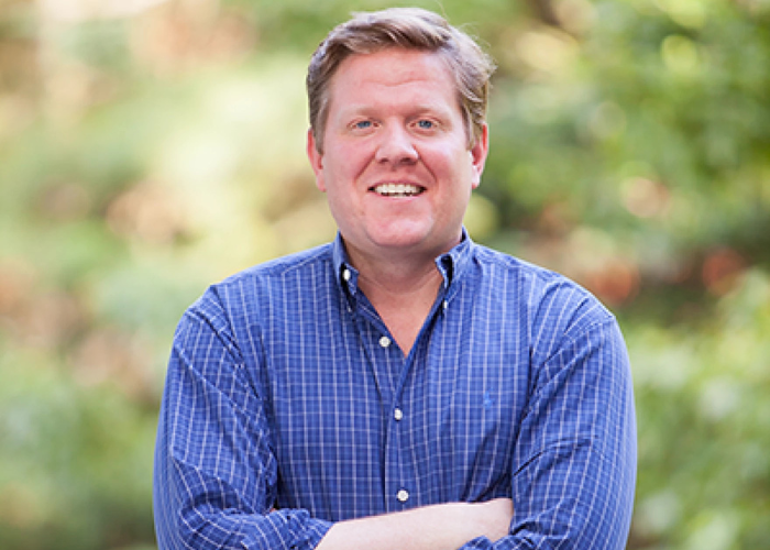 A smiling man in a blue button-down shirt with his arms folded.