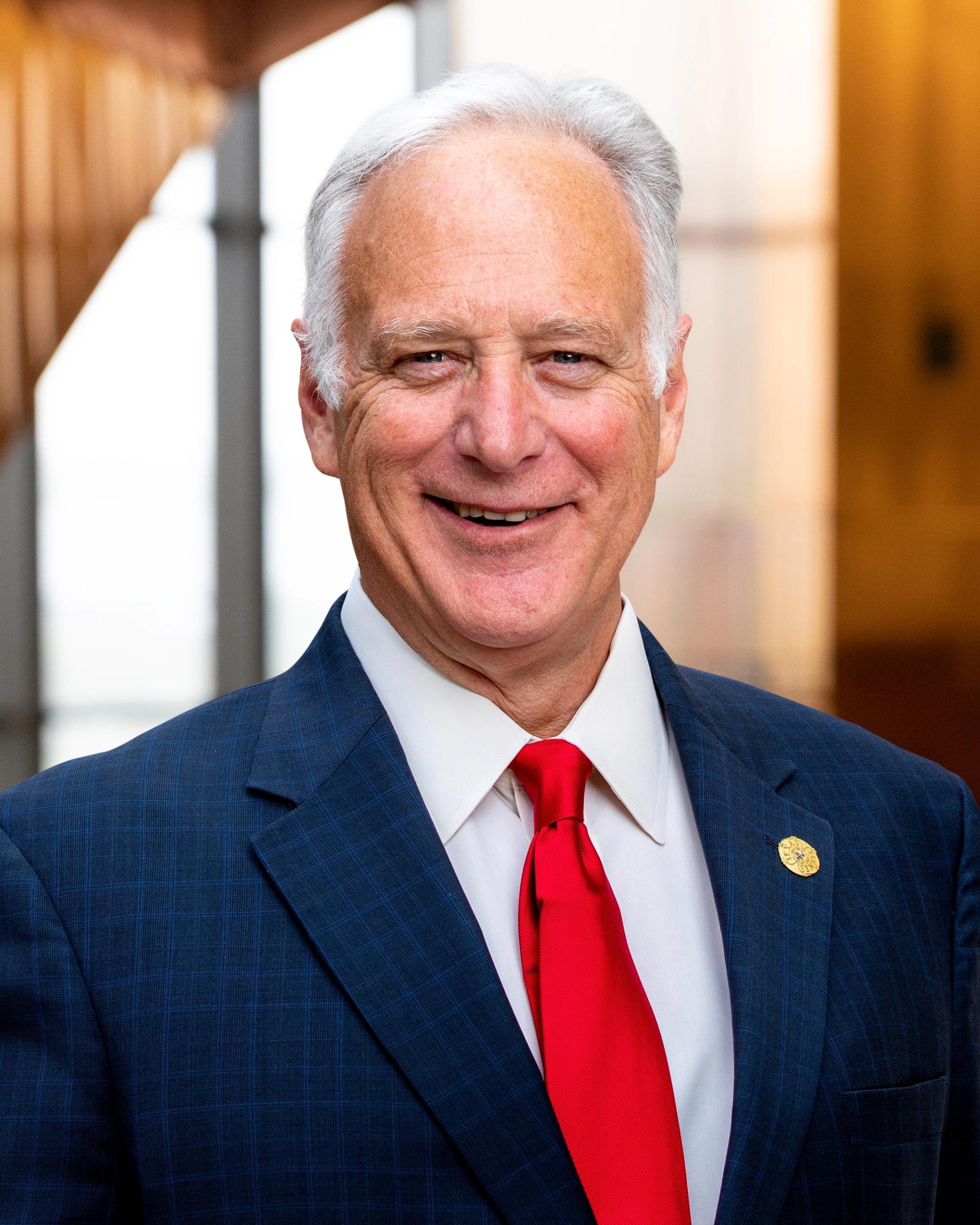 Kirk Watson smiling in a blue suit, white shirt, and red tie.