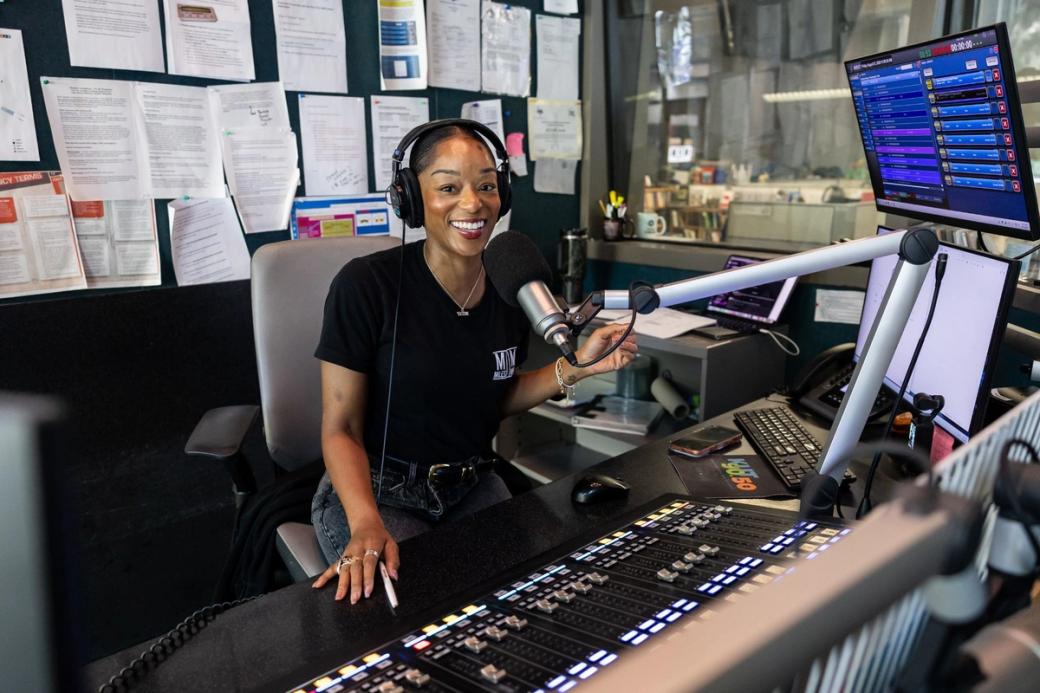 A woman wearing a t-shirt behind a microphone in a radio studio.