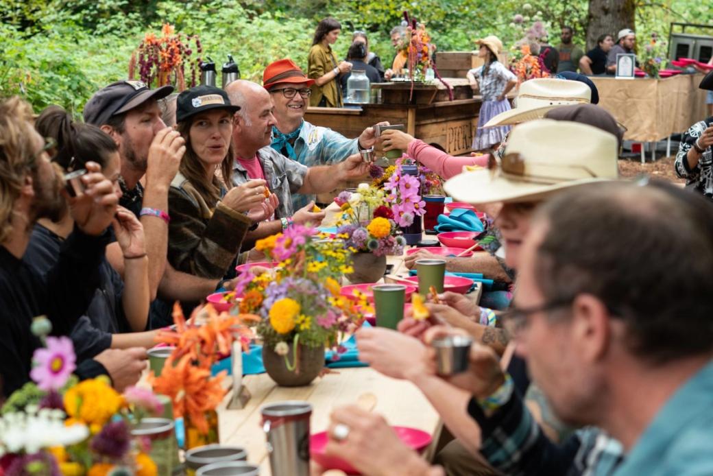 A group of people enjoying dress at a long table outside at an event decorated with flowers.