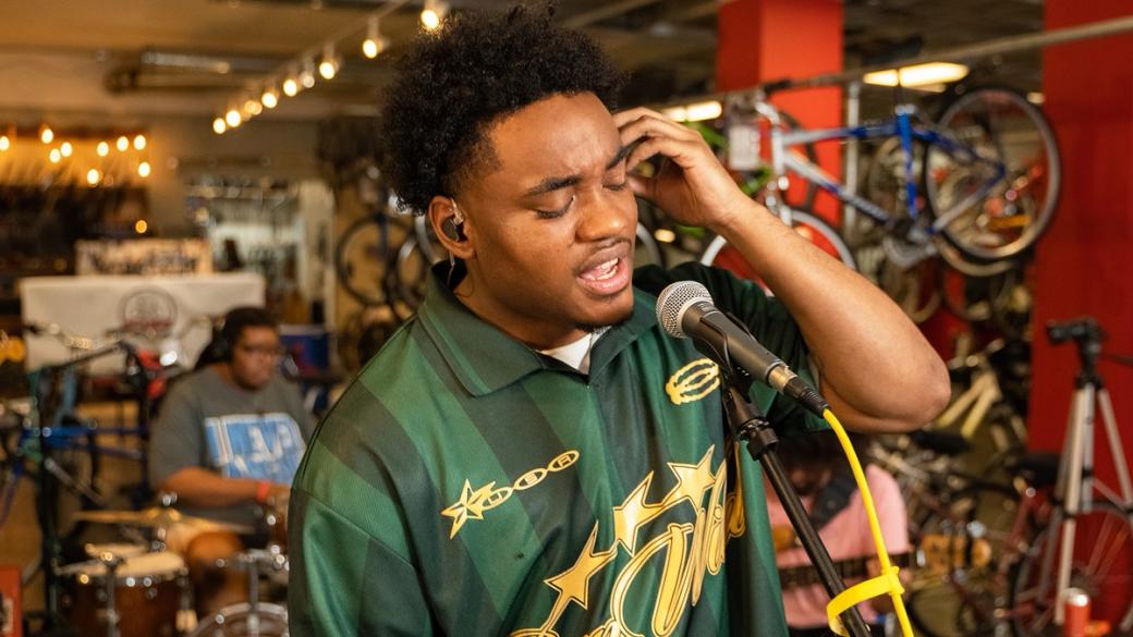 Man sings into microphone inside bicycle shop, with bikes and drummer in background