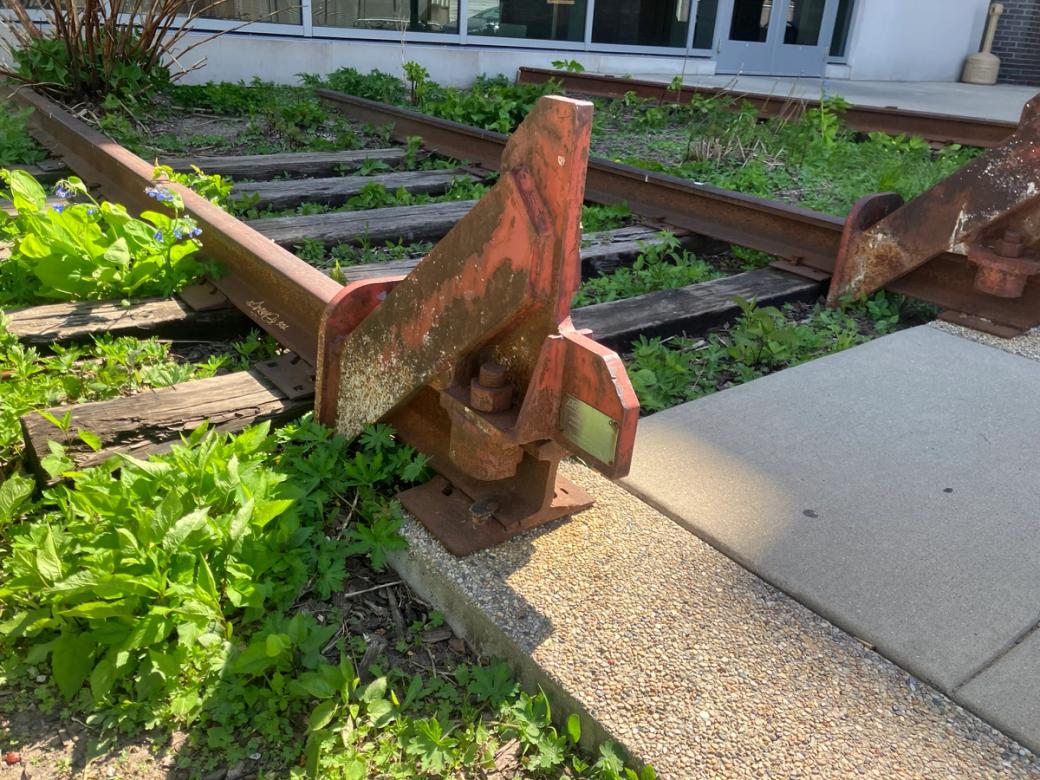 A short, rusty section of train track next to a sidewalk. The track is covered in grasses and plants, and is right next to a white building.