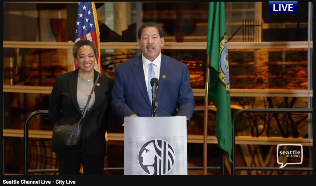 Seattle Mayor Bruce Harrell delivers his concession speech with his wife, Joanne Harrell at a podium next to an American and Seattle flag.