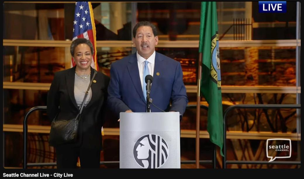 Seattle Mayor Bruce Harrell delivers his concession speech with his wife, Joanne Harrell at a podium next to an American and Seattle flag.
