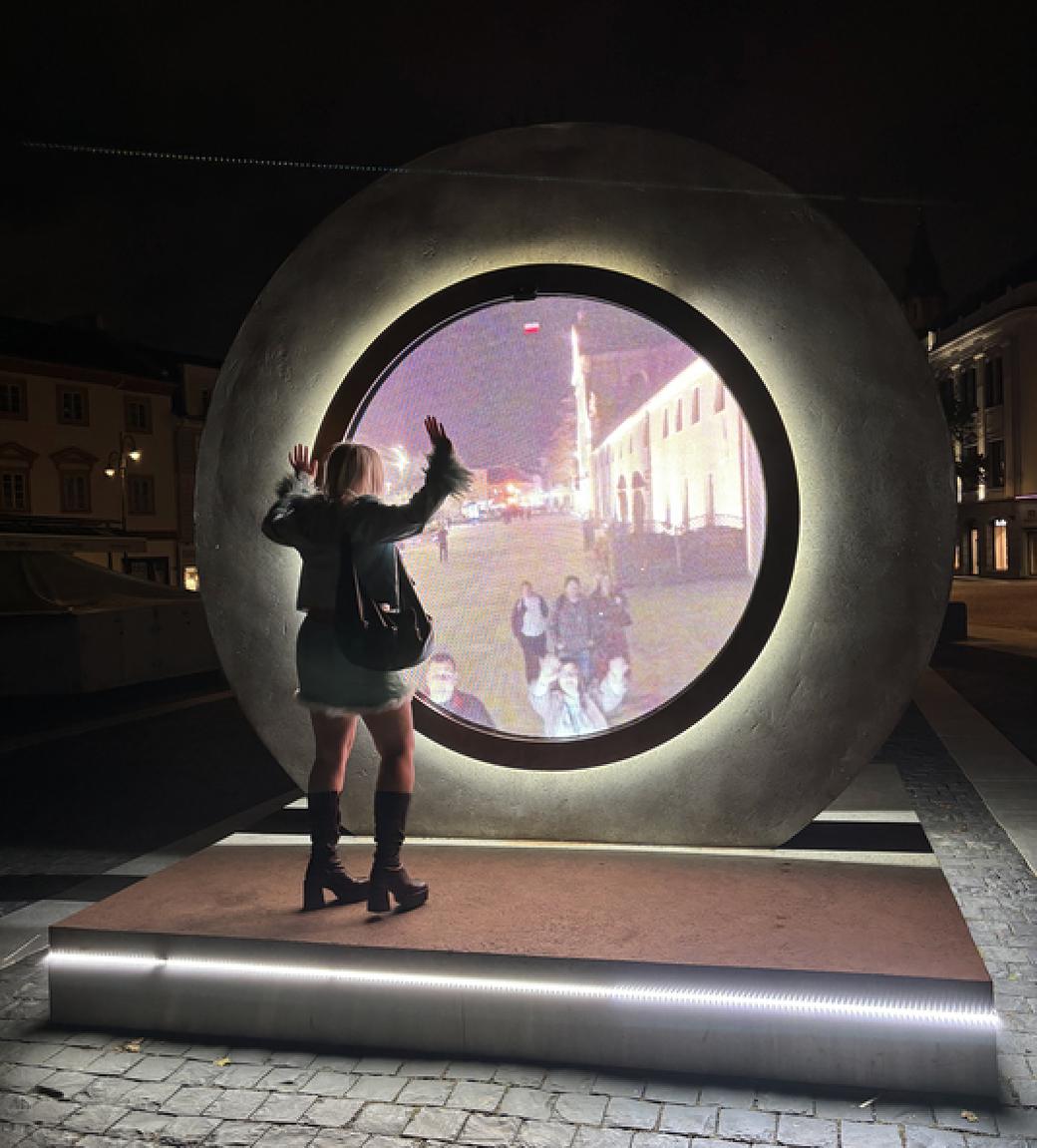 A woman standing on a platform in front of a large circular piece of stone. In the middle of the stone, there's a circular screen with a live stream of other people at another location