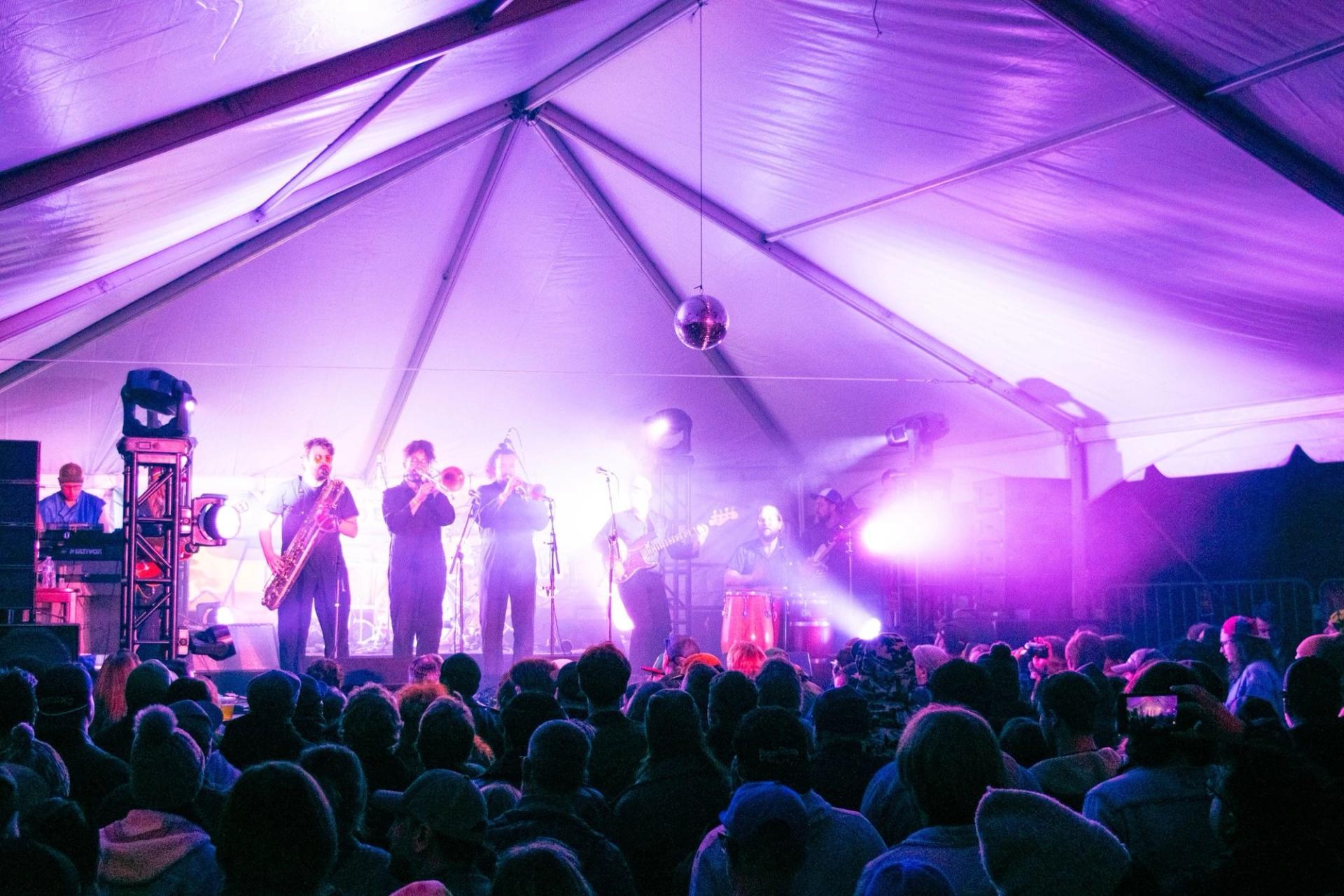 A band playing for a crowd inside of a tent with purple lighting