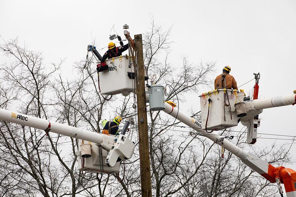 Booms lift lineworkers to electric poles surrounded by trees in winter.