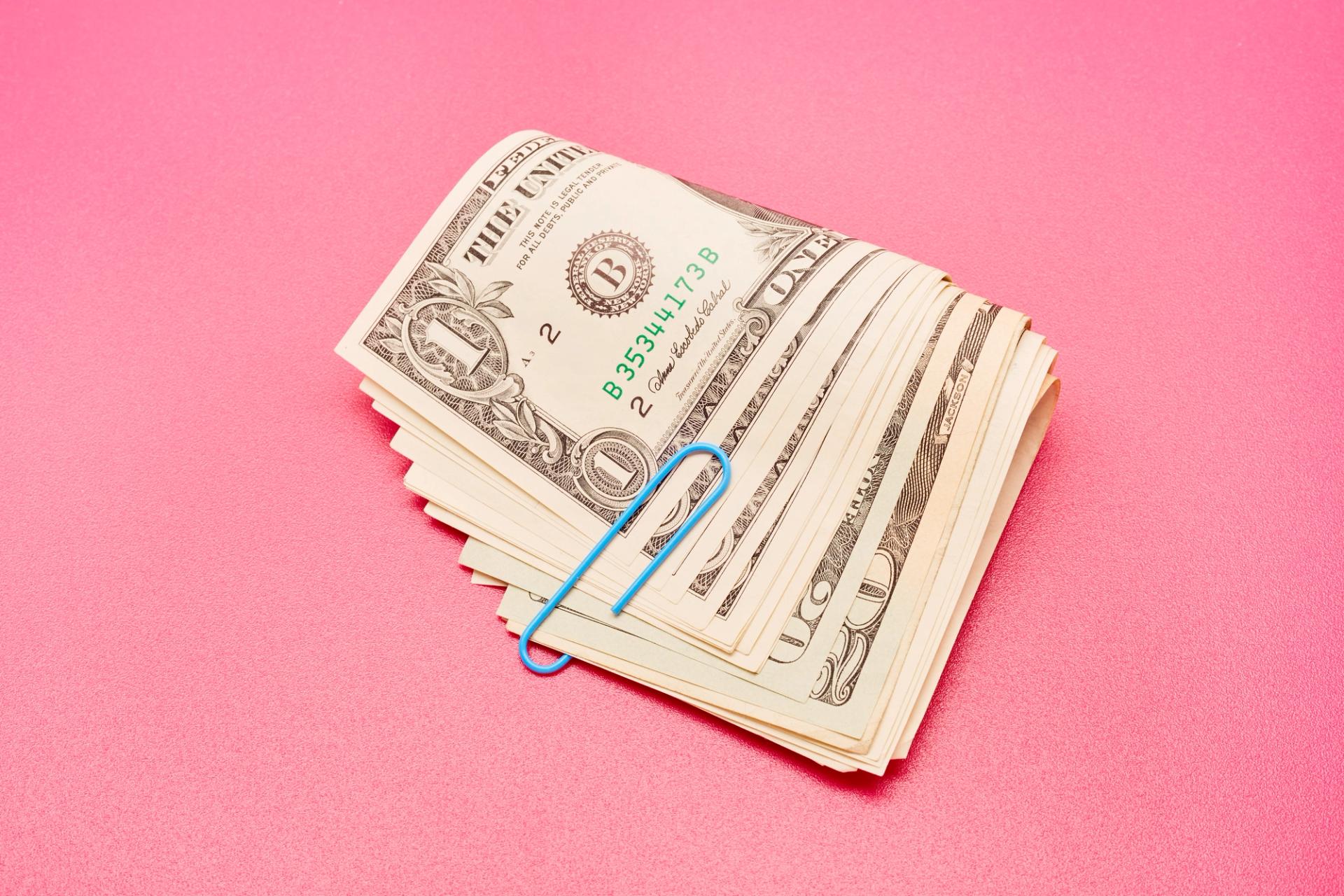 A wad of cash held together with a blue paper clip on a pink background.