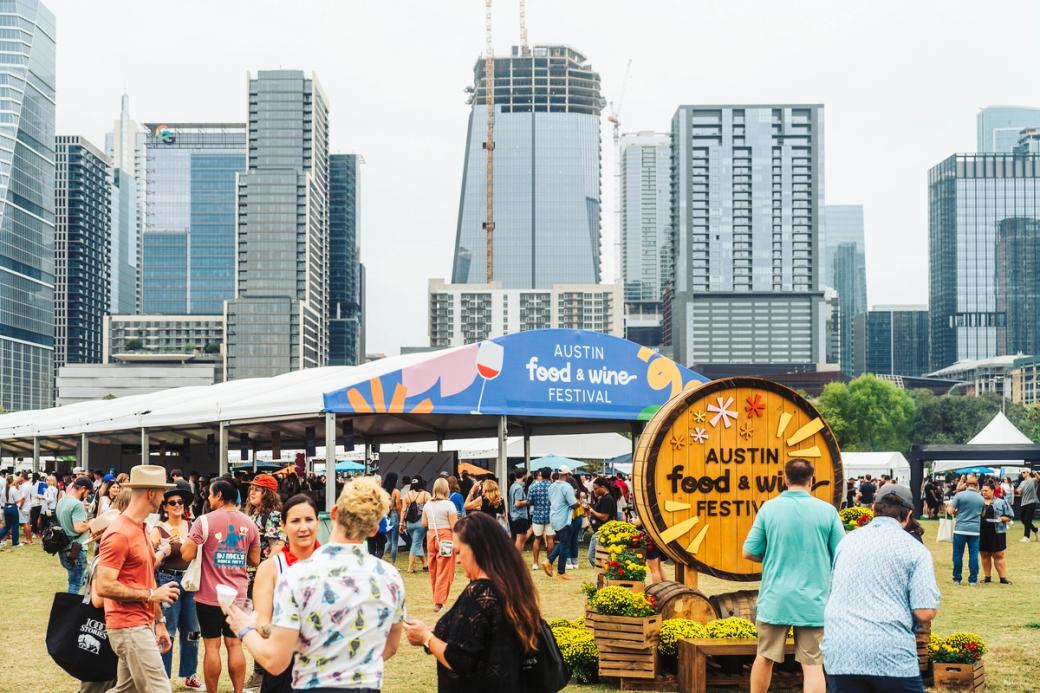 A bunch of people standing in a field with the Austin skyline in the background. A tent that says "Austin Food & Wine Festival" is up.