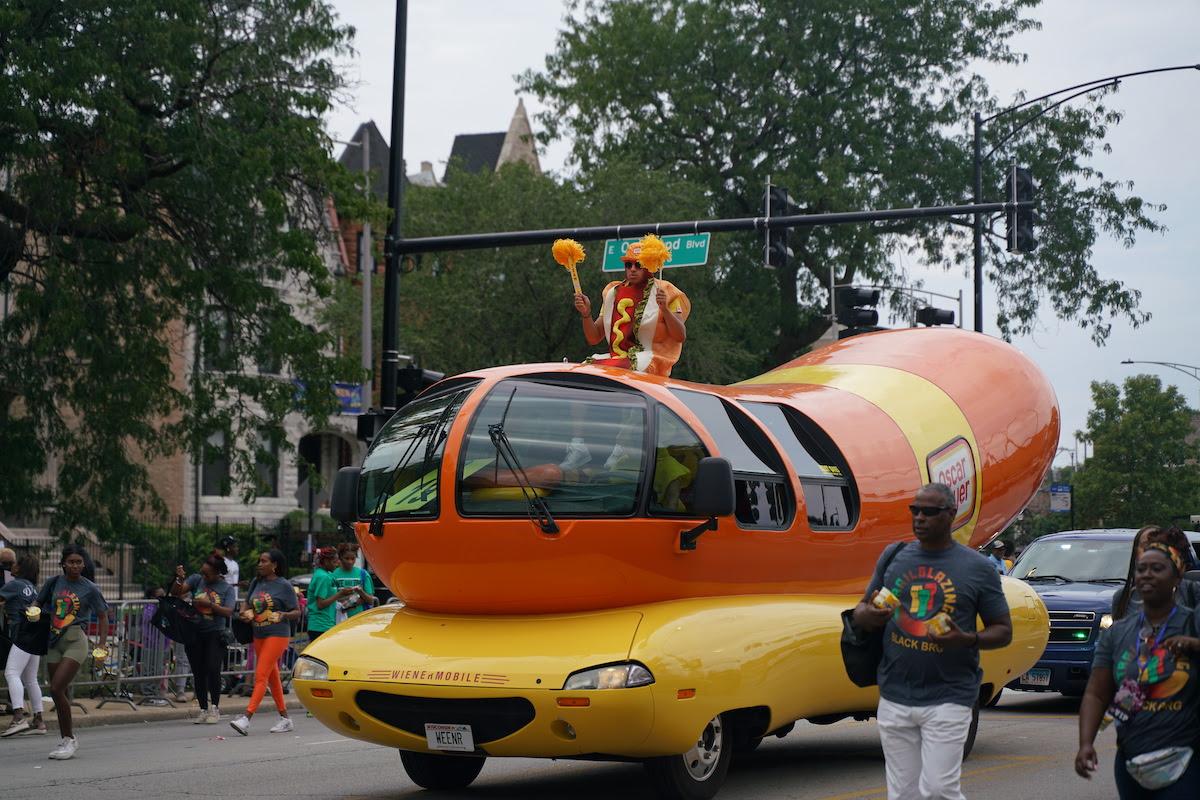 A person dressed as a hot dog rides in a hot dog vehicle in a parade