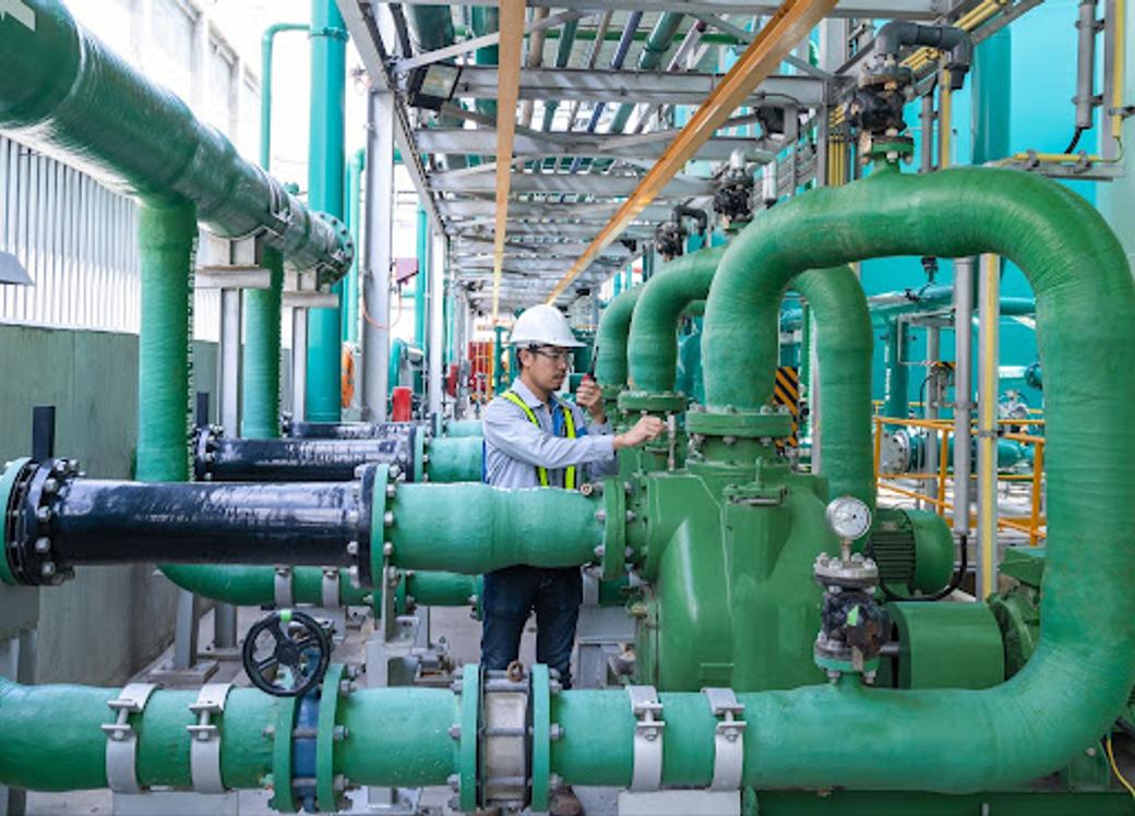 An engineer wearing a hard hat and safety vest adjusts equipment among large green pipes and pumps and water processing facility. 