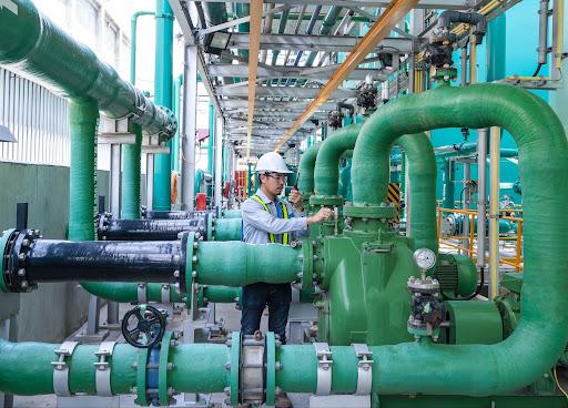 An engineer wearing a hard hat and safety vest adjusts equipment among large green pipes and pumps and water processing facility.