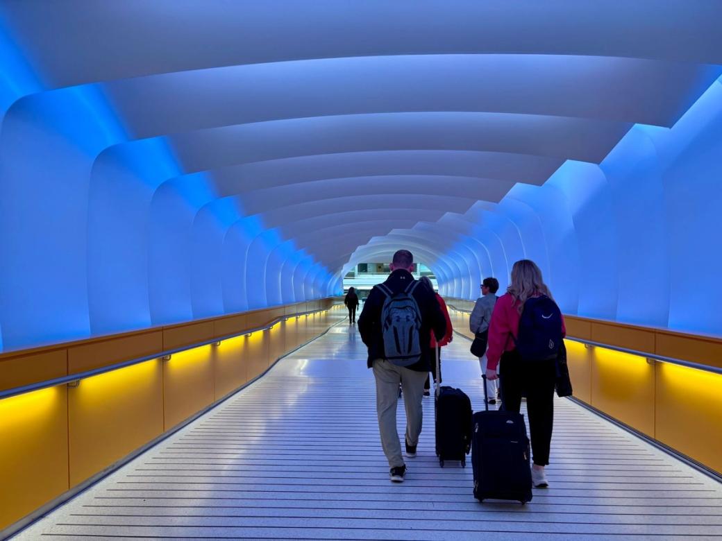 People walk with their luggage down a colorfully lit tunnel.