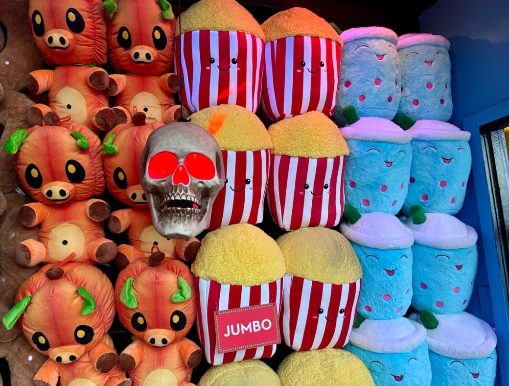 A display packed with oversized plush prizes, including orange pig-like characters with pumpkin heads, smiling red-and-white popcorn buckets, and blue drinks, with a glowing red-eye skull hanging in the center.