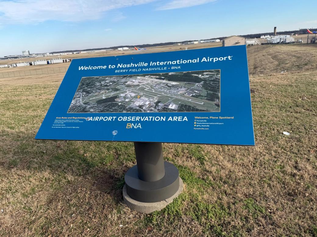 A blue sign in front of a field, with planes and trucks in the far distance. The sign says: "Welcome to Nashville International Airport." "Airport Observation Area BNA."