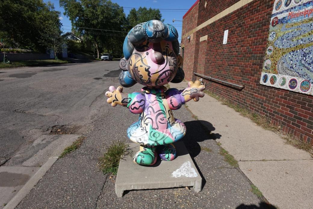 A colorful statue of the Lucy character from Peanuts sits on a road next to a red brick building.