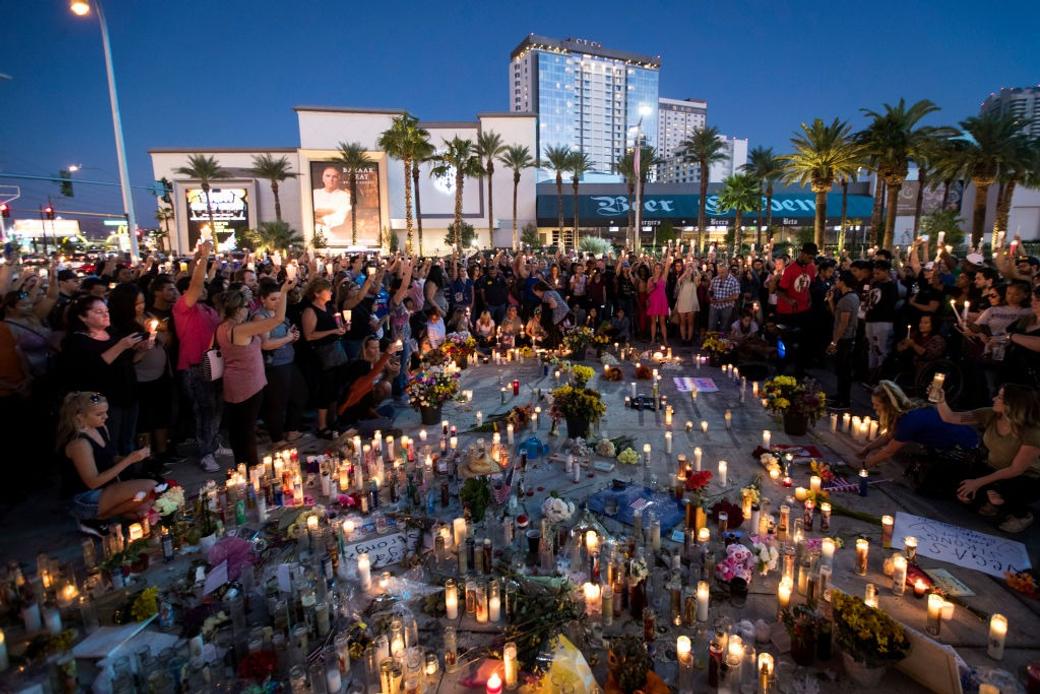 Crowd holds candles at a nighttime Las Vegas memorial vigil surrounded by flowers, candles, and a "Vegas Strong" sign on the ground.