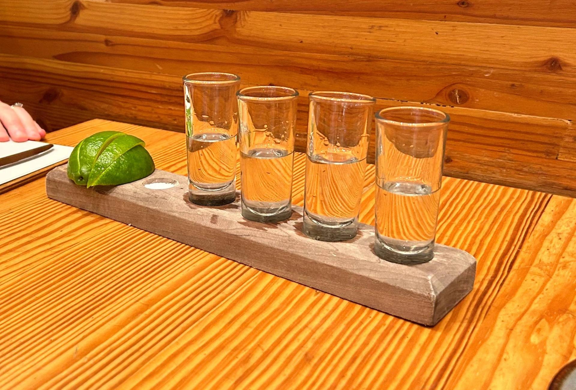 Four glass shot glasses filled with clear tequila sit on a wooden plate.