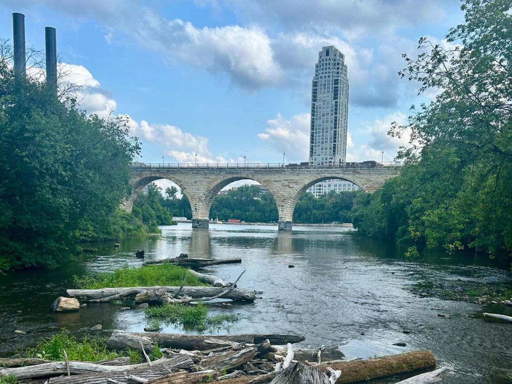 Eye-level view of river with brick bridge going over it, trees on the water's edge, and large building in the distance