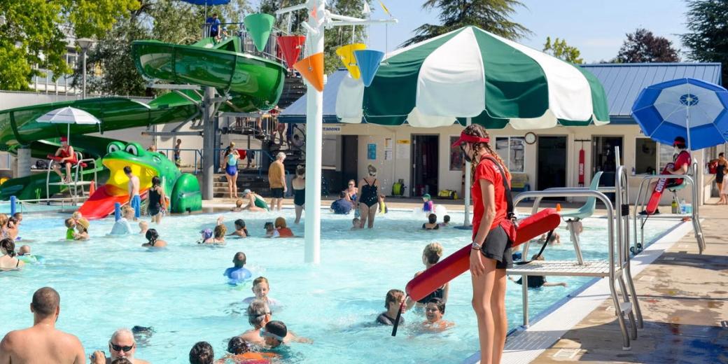 people in a pool, with a lifeguard on the edge. In the background is a fog shaped slide into the pool for kids.