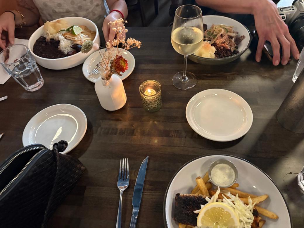 A brown wooden table with multiple white plates, three of which have food on them. There's also wine and water glasses, and a small white vase with dried flowers. A candle lights the table.