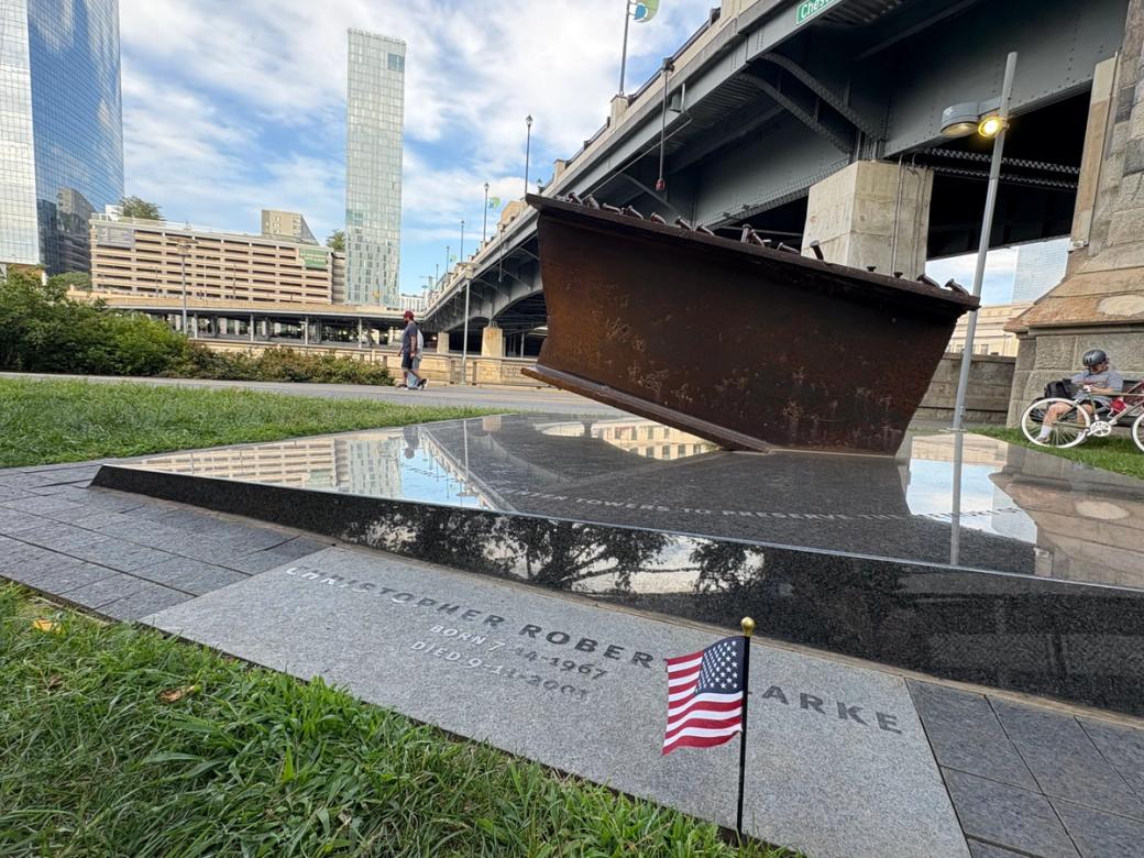 A piece of a rusted steel beam from the World Trade Center is displayed on a marble podium along the Schuylkill River Walk. At the foreground is an engraving honoring Christopher Robert Clarke, born 7-14-1967 and died 9-11-2001. A small American flag is propped up on the grass.