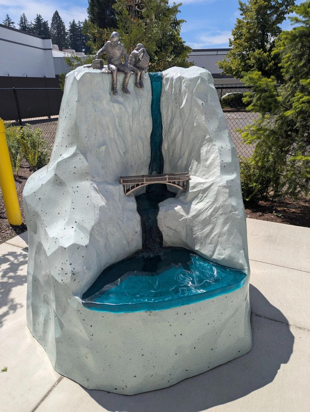 A blue, white, and steel sculpture of two people having a picnic at the top of a waterfall.
