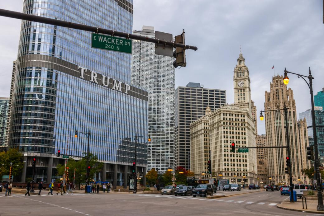 An intersection with people walking. The street says "E Wacker Dr" There is a large glass building that says "TRUMP" on it