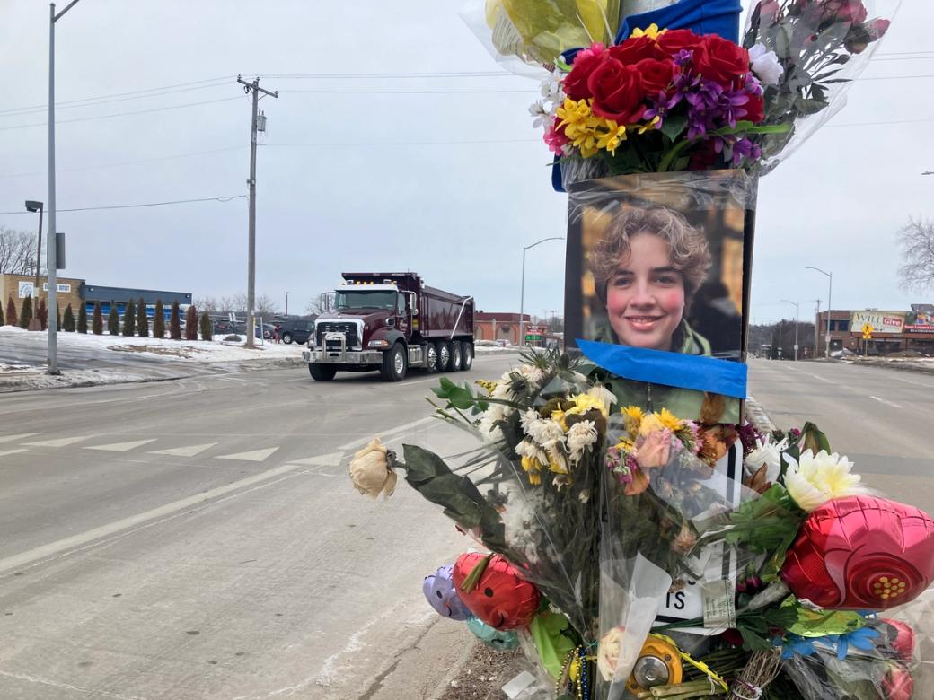 A roadside memorial on South Park Street with flowers, balloons, and a photo of Sasha Rosen. The street is mostly empty, with one truck in the background.
