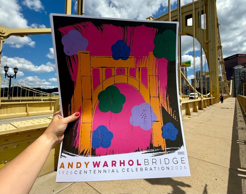 A colorful poster reading "ANDY WARHOL BRIDGE" leaning on a yellow railing over a river.