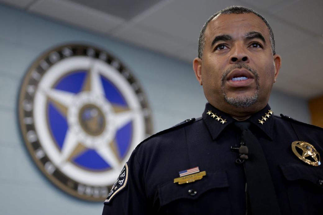 A close-up of a uniformed Black man, with the circular, multi-point star seal of the Denver Sheriff Department on the wall behind him.