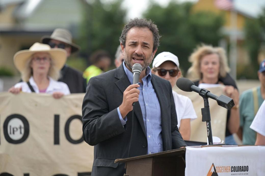 A man in a suit holding a microphone stands at a podium that says "Together Colorado." Behind him, several people are holding a sign with a message that's not fully visible in this photo.