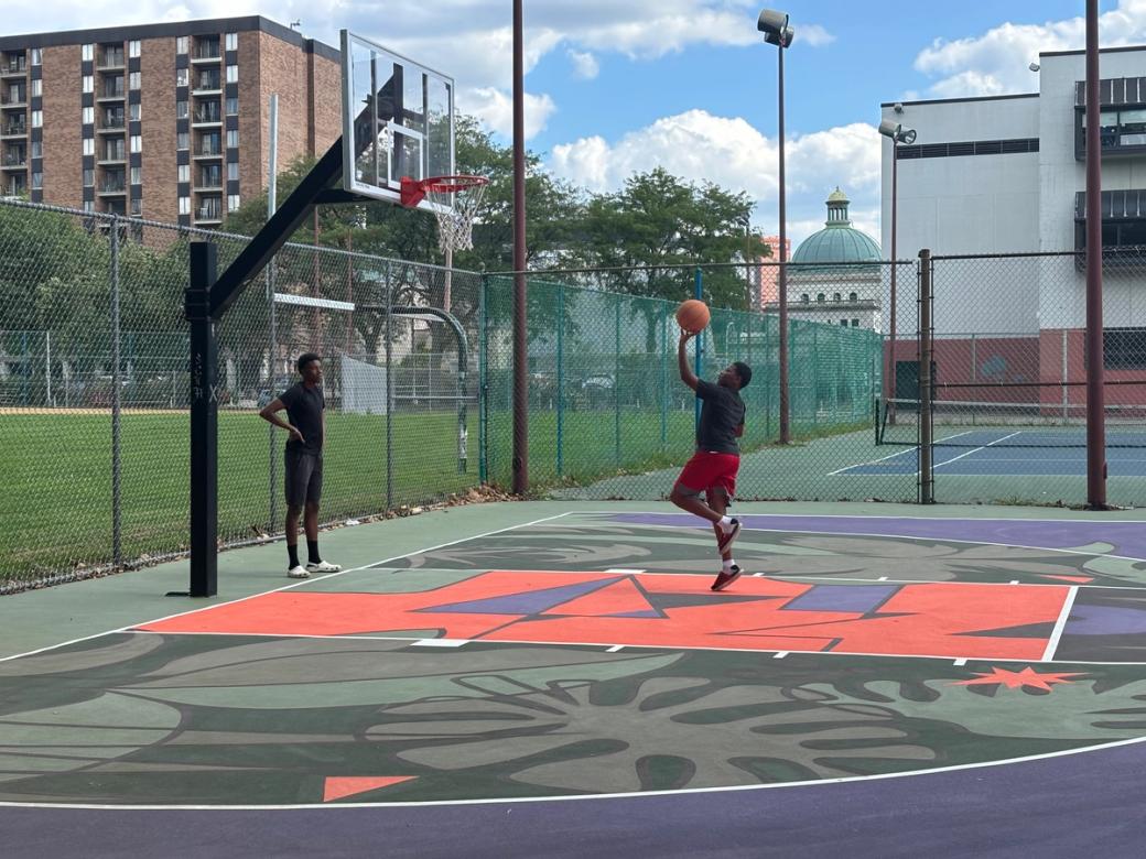 Two Black youth playing basketball on the newly renovated court designed with plant and music symbols in orange, green, and purple