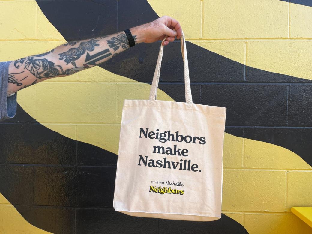 A tattooed arm holds the City Cast Nashville canvas tote in front of a brick wall with wavy yellow and black stripes. The tote says Neighbors make Nashville. City Cast Nashville Neighbors in yellow and black lettering.