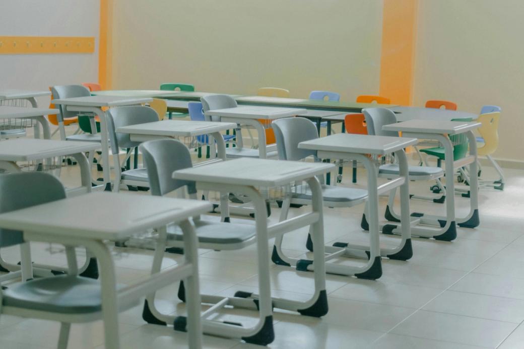 Empty gray chairs sit in a classroom meant for young students. Some colorful chairs sit in a semicircle towards the back of the image, with yellow accents on the wall behind them.