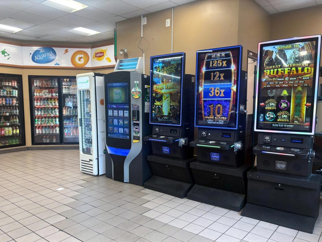 Three large skill game consoles are set up next to each other in the brightly lit market of a gas station. One screen reads "Great American Buffalo," and another reads "Plunderin' Pirates," all of which promise a chance to win money. To the left of these games, fridges are full of soft drinks and other beverages.